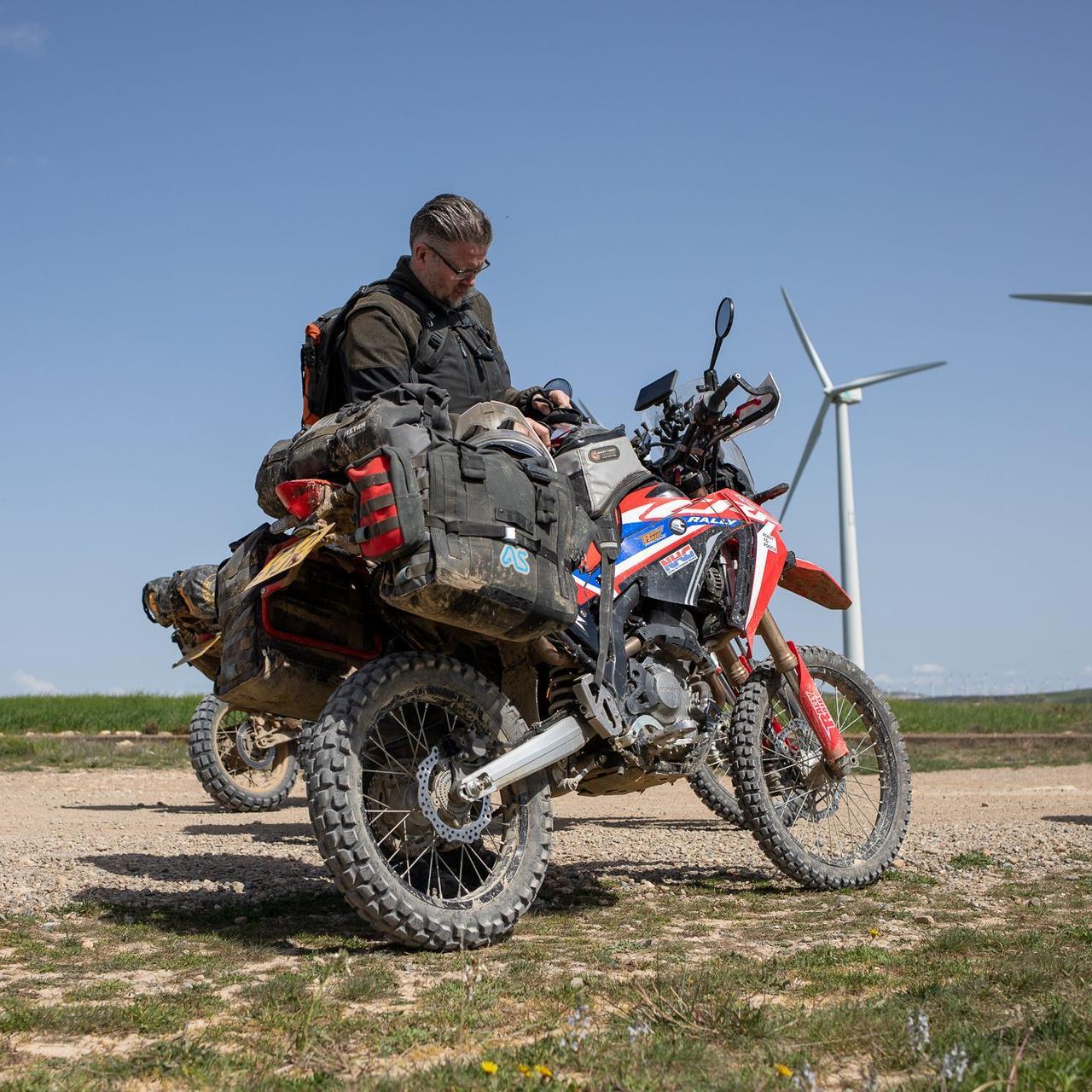 rider on dusty trail standing behind honda crf300 rally motorcycle with magadan panniers mounted in foreground