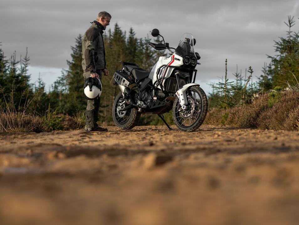 man in grey gravel jacket and pant holding white helmet looking at white ducati desert x motorbike with adventure spec hard parts fitted